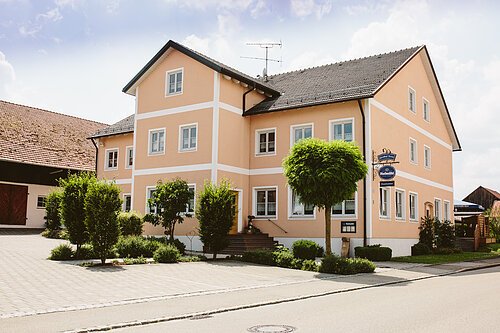Der Gasthof von außen von der Straße aus eingefangen. Der Gasthof ist in einem schönem Ockerton gestrichen. Die Fenster und Türen sind mit weiß umrandet. Links ist der gepflasterte Hof mit Scheune.