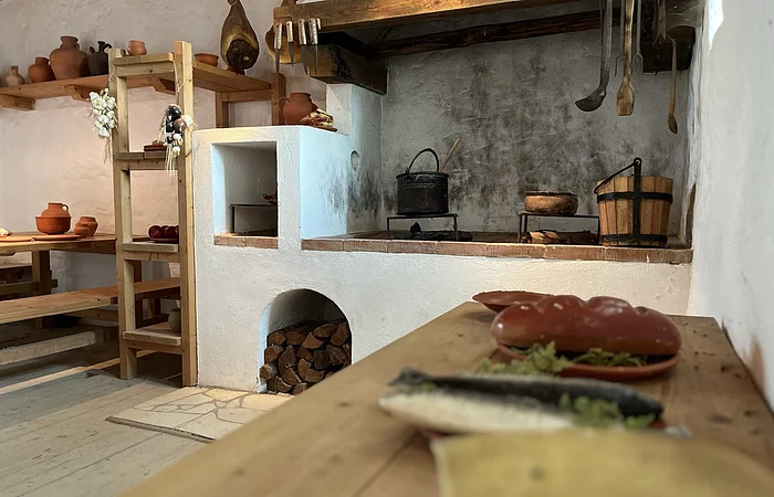 Der große offene Herd mit zwei Töpfen nimmt viel Raum ein. Links sind gefüllte Regale zu sehen. Im Vordergrund ist eine hölzerne Arbeitsplatte auf der Teller stehen.