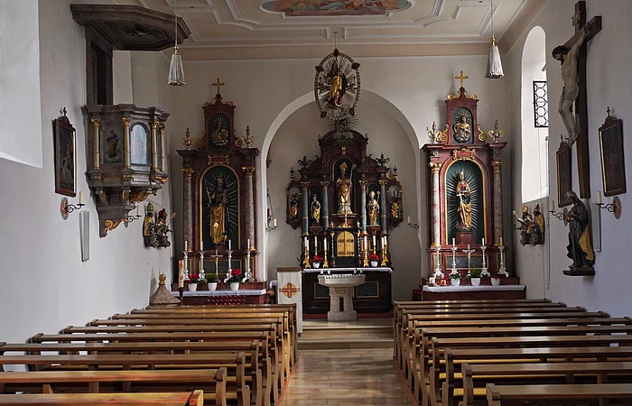 Innenraum einer Kirche mit Holzbänken, Altären, religiösen Statuen und einem Kruzifix an der rechten Wand.