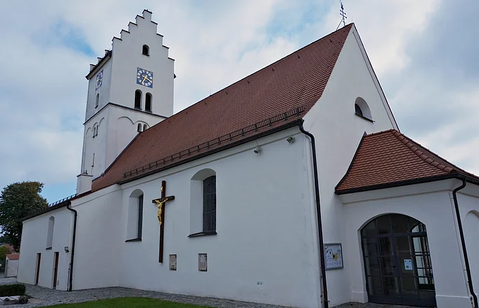 Weiße Kirche mit rotem Dach, Uhrturm und großem Kreuz an der Außenwand bei bewölktem Himmel.